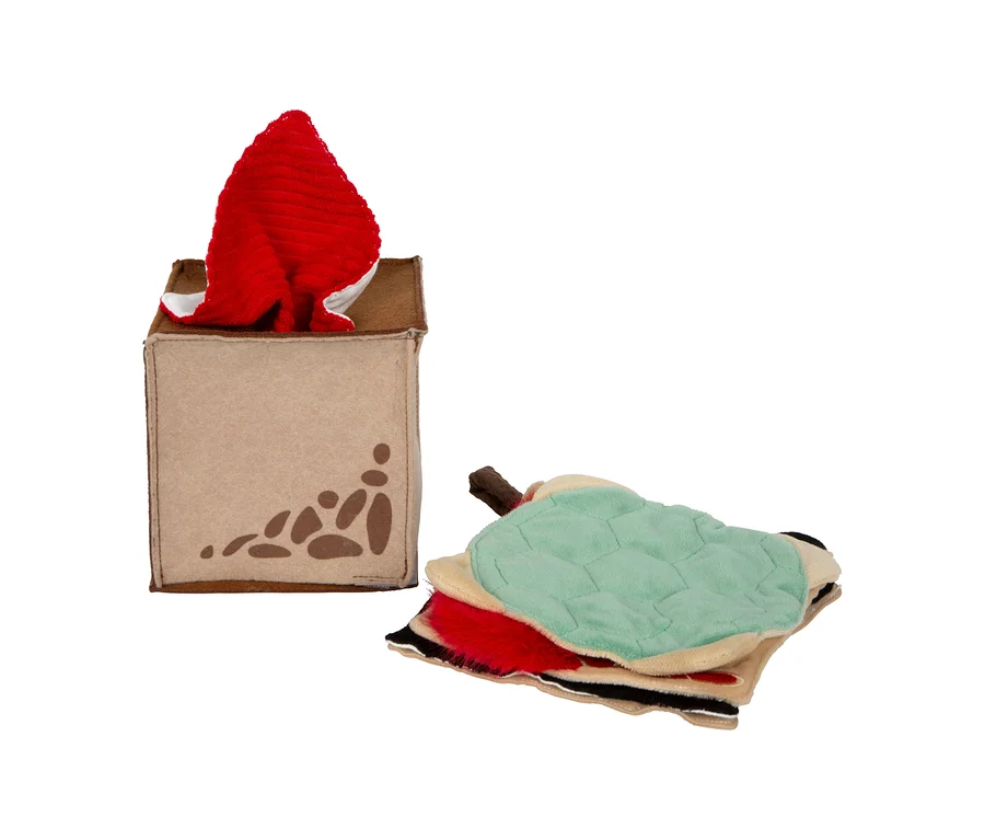 Eine beigefarbene small foot Baby-Box, 7–9 Monate, mit braunen Mustern, aus der ein rotes Plüschtier ragt. Daneben liegt ein Stapel bunter Stofftiere mit grünem Oberteil.