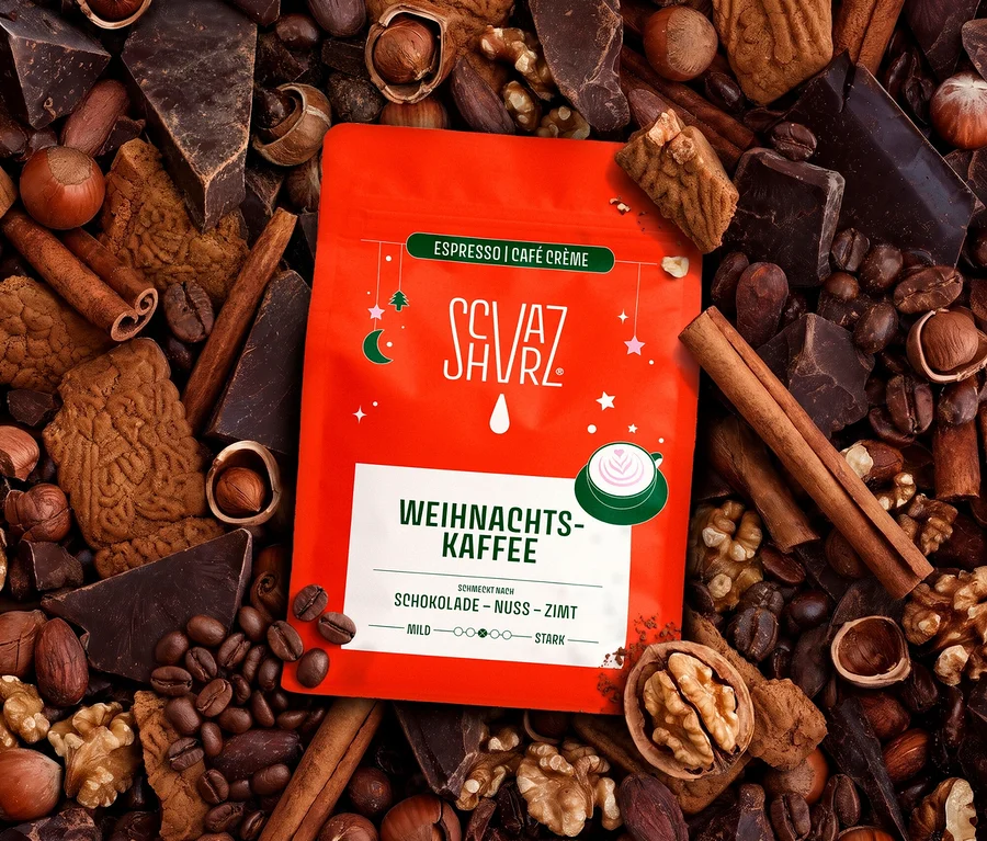 Ein roter Beutel Schvarz - Weihnachtskaffee Espresso liegt inmitten von zerbrochener dunkler Schokolade, ganzen Haselnüssen, Walnüssen, Kaffeebohnen, Zimtstangen und Spekulatius-Keksen. Der Beutel hat weißen Text und ein grünes Etikett mit "Weihnachtskaffee" und den Geschmacksrichtungen Schokolade, Nuss, Zimt.