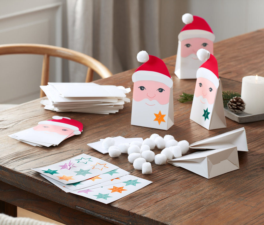 Auf einem Holztisch liegt ein DIY-Adventskalender-Set mit Santa-Gesicht-Papiertüten, Sternaufklebern, Wattebällchen und Kerzen.
