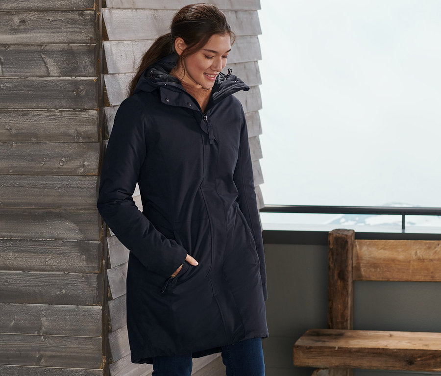 Eine Frau steht an einer Holzwand mit Skiern und trägt einen blauen Thermo-Mantel.