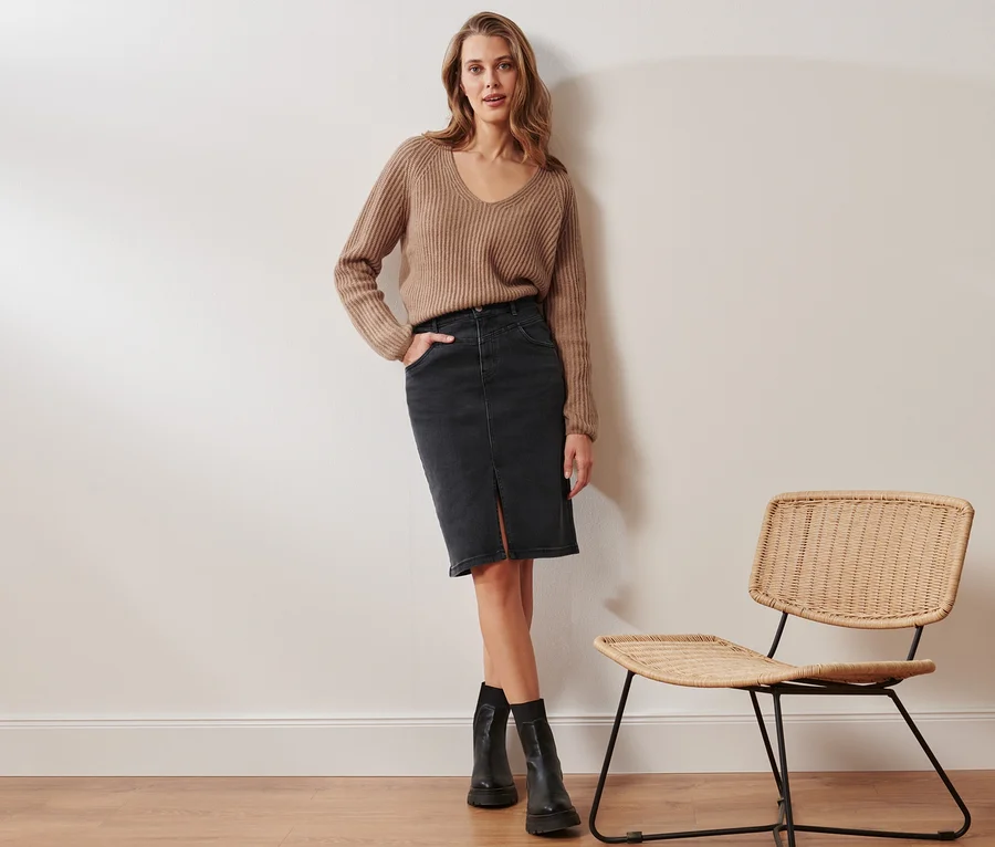 Eine Frau posiert in einem beigen Grobstrick-Pullover mit Wolle und einem Jeansrock in Bleistift-Form. Neben ihr steht ein Rattanstuhl.