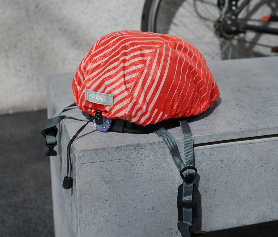 Ein Fahrradhelm-Regenüberzug mit orangen Streifen liegt auf einer Betonoberfläche, im Hintergrund ein Fahrrad.
