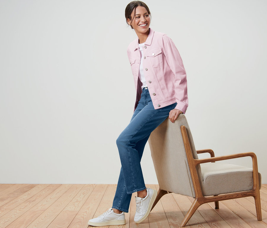 Lächelnde Frau posiert in rosa Jeansjacke und Jeans an einem Sessel.