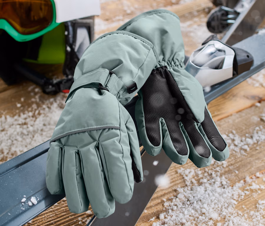 Grüne Kinder-Ski- und Winterhandschuhe liegen auf Skiern neben Helm und Brille.