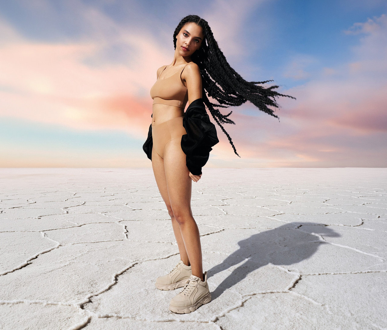 Frau mit Zöpfen trägt nostalgisch-braunes sloggi ZERO Feel 2.0 Top und eine offene, schwarze Jacke vor einem hellen Hintergrund.