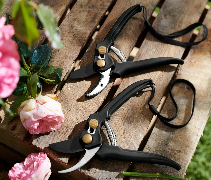 Zwei schwarze Gartenscheren liegen auf einer Holzpalette neben rosa Rosen. Gartenscheren-Set.