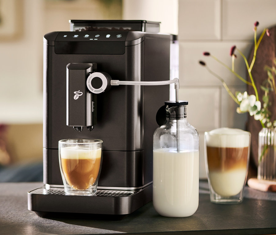 Ein »Esperto 2 Milk« Granit Black Kaffeevollautomat bereitet Kaffee in einem Glas zu. Daneben steht eine Milchkaraffe.