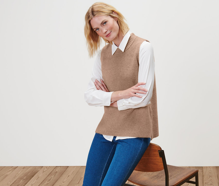 Eine blonde Frau posiert auf einem Stuhl in einem beige melierten Strick-Pullunder, einem weißen Hemd und blauen Jeans.