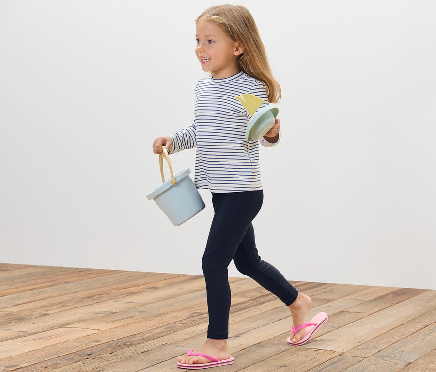 Ein Mädchen geht auf einem Holzboden und trägt einen blauen Eimer und Sandspielzeug. Sie trägt eine Kinder-Badeleggings mit UV-Schutzfaktor 80.