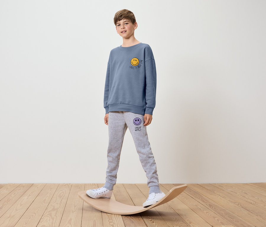 Ein Junge in einem blauen Smiley-Print-Sweatshirt und grauer Jogginghose steht auf einem Balance Board.