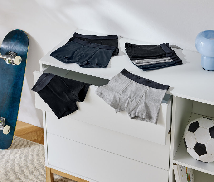 Auf einer Kommode liegen Herrenboxershorts, daneben stehen ein Skateboard und ein Fußball.
