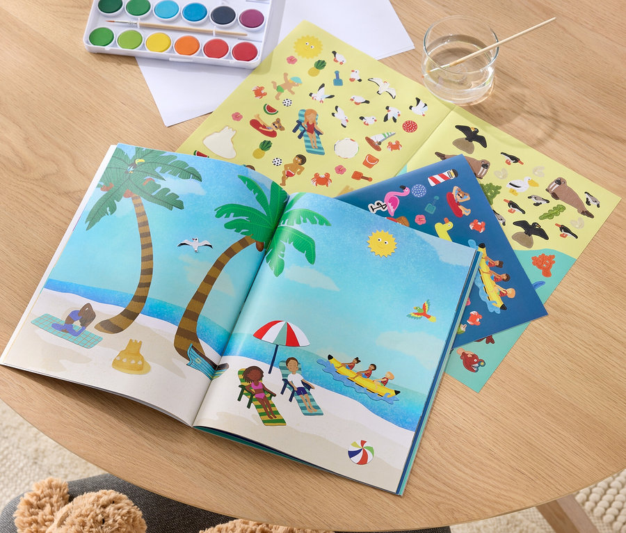 Ein geöffnetes Mein Stickerbuch »Am Meer« mit Strandillustrationen, Farben zum Malen und Stickerbögen auf einem Holztisch.
