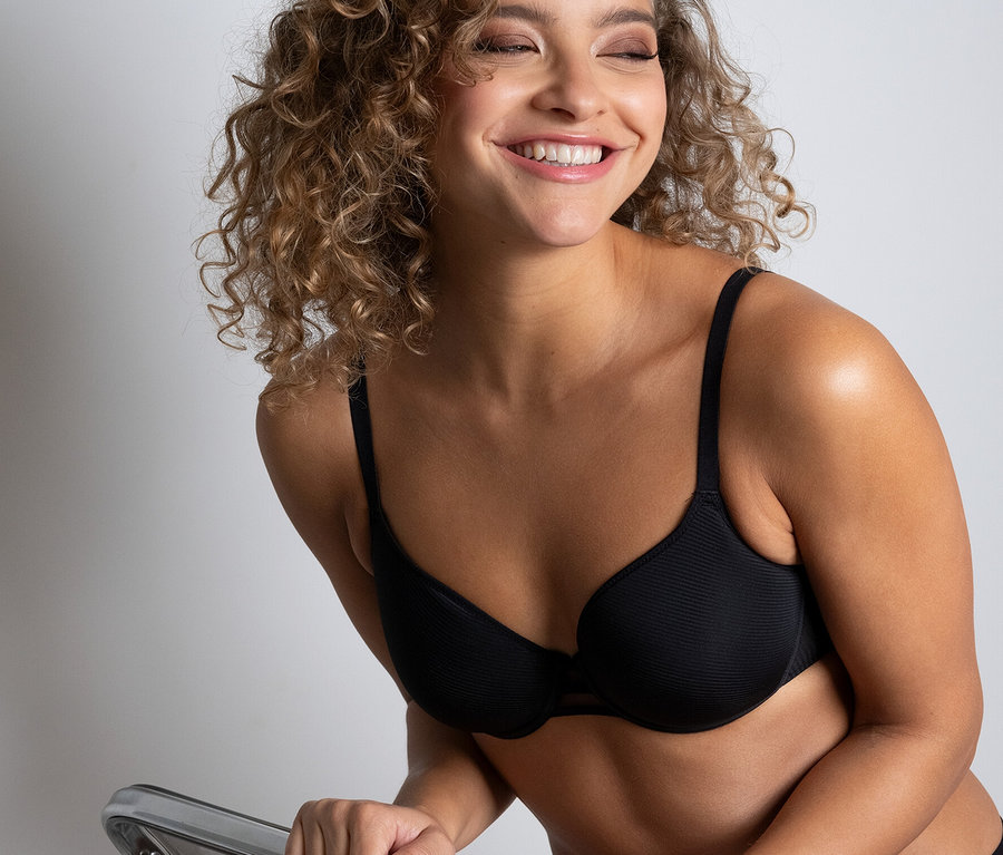 Lächelnde Frau sitzt auf einem durchsichtigen Stuhl und trägt einen schwarzen Passionata Spacer-BH »Nais«.
