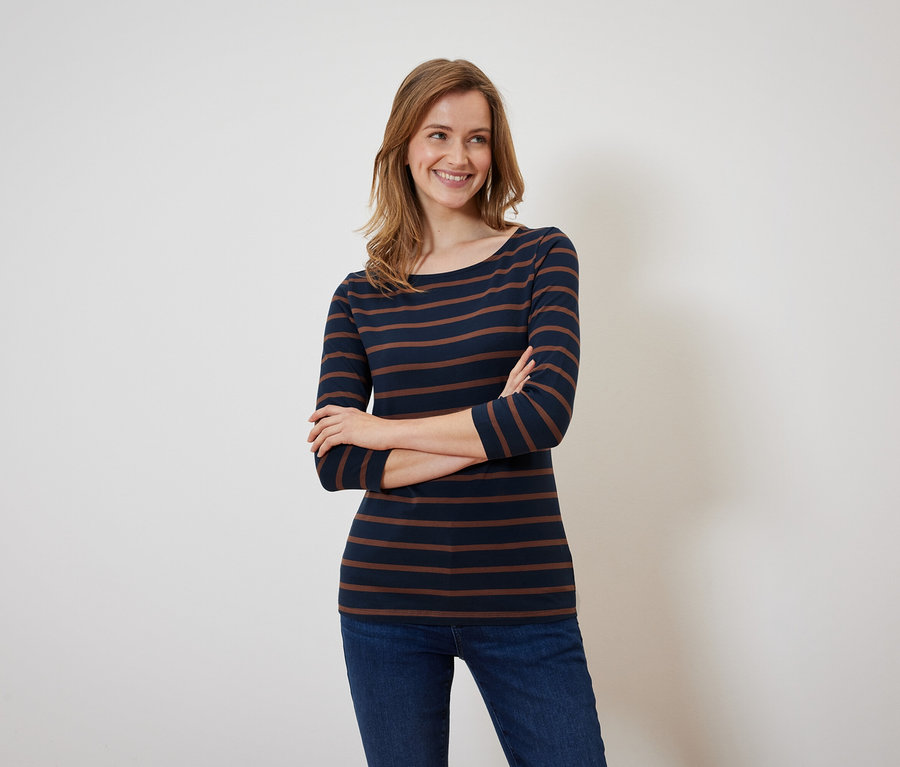 Eine lächelnde Frau posiert mit verschränkten Armen in einem marineblau-braun gestreiften Shirt und Jeans.