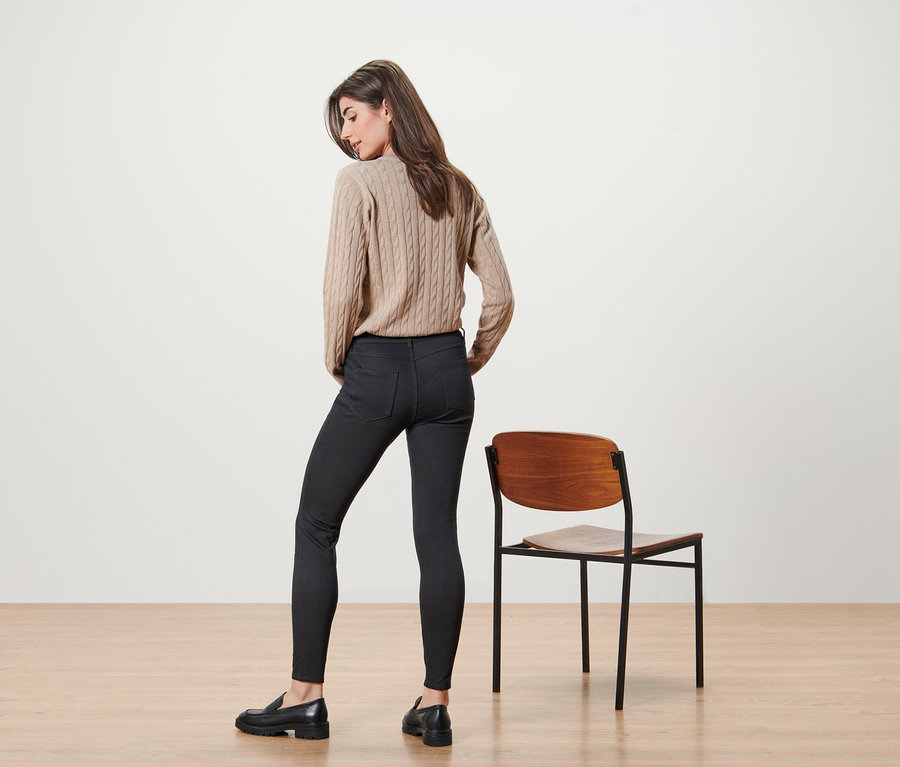 Frau posiert in beigem Pullover und schwarzen Jeans neben einem Stuhl.