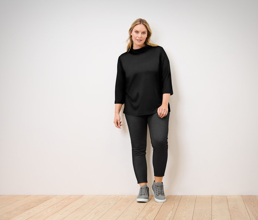 Frau posiert in einem schwarzen Oberteil mit Dreiviertelärmeln, schwarzen Jeans und grauen Turnschuhen.