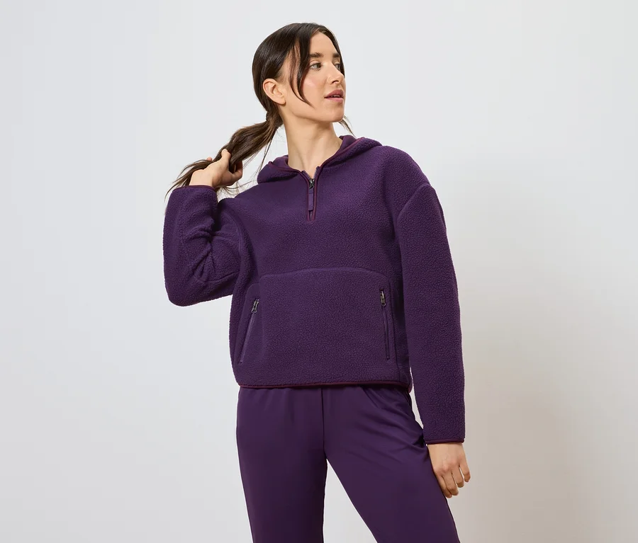 Frau hält Zopf und trägt einen lila Kapuzenpullover und Hose.