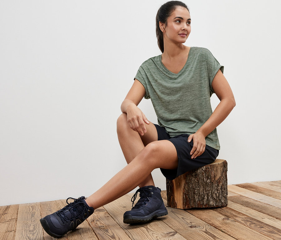 Frau sitzt auf einem Holzscheit und trägt ein khaki Funktionsshirt.