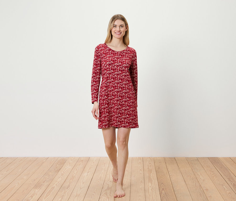 Eine Frau geht barfuß über einen Holzboden und trägt ein rotes, langärmliges Kleid.