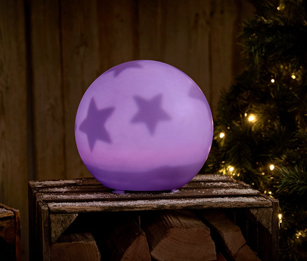 Lila Kugel mit Sternen auf einer Holzkiste neben einem Weihnachtsbaum.