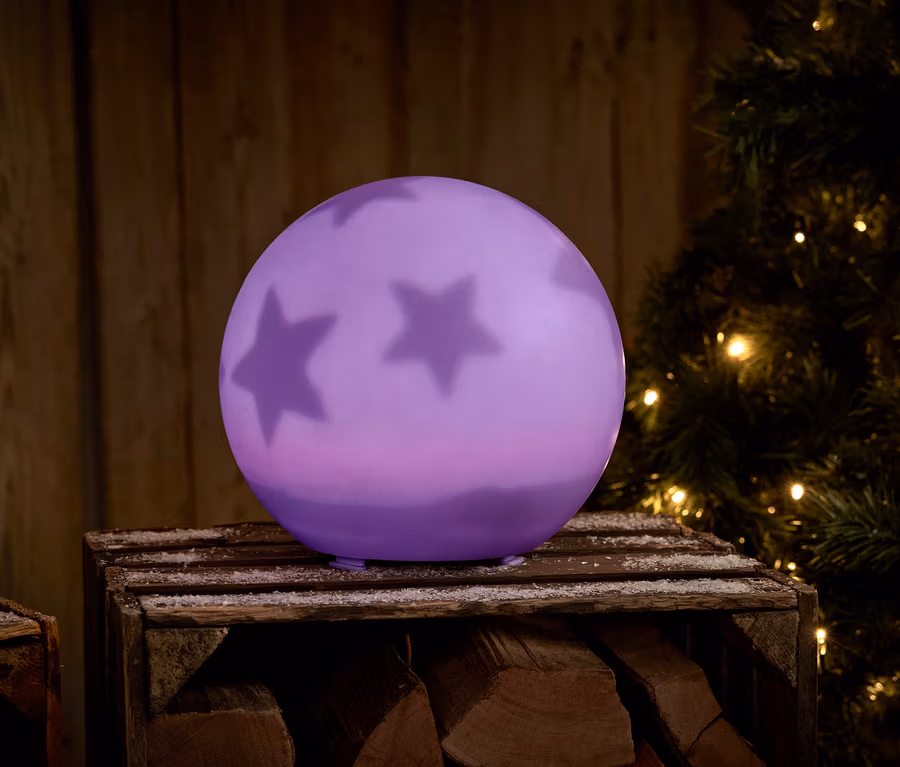 Lila Kugel mit Sternen auf einer Holzkiste neben einem Weihnachtsbaum.