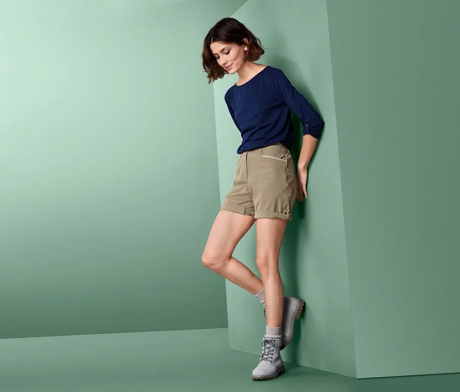 Eine Frau, die sich an eine Wand lehnt, trägt Softshell-Funktionsshorts, ein blaues T-Shirt und graue Stiefel.