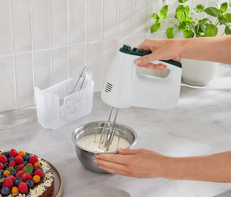Frau mixt Teig in einer Schüssel mit einem Handmixer neben einem Kuchen mit Früchten.