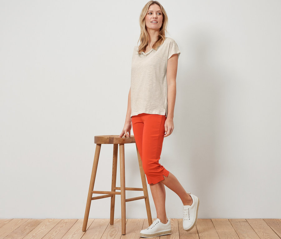 Blonde Frau posiert neben einem Holzhocker in orangefarbener 3/4-Stretchhose.