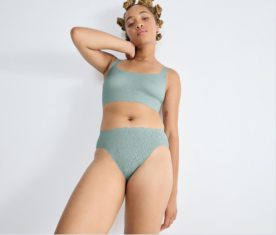 Eine Frau mit blonden Dreadlocks steht vor einer weißen Wand und trägt einen misty turquoise sloggi ZERO Feel Bliss High leg Slip und ein passendes Bralette. Sie hat einen Arm hinter dem Kopf erhoben und blickt in die Kamera. Beide Teile haben ein dezentes Muster und gewellte Kanten.