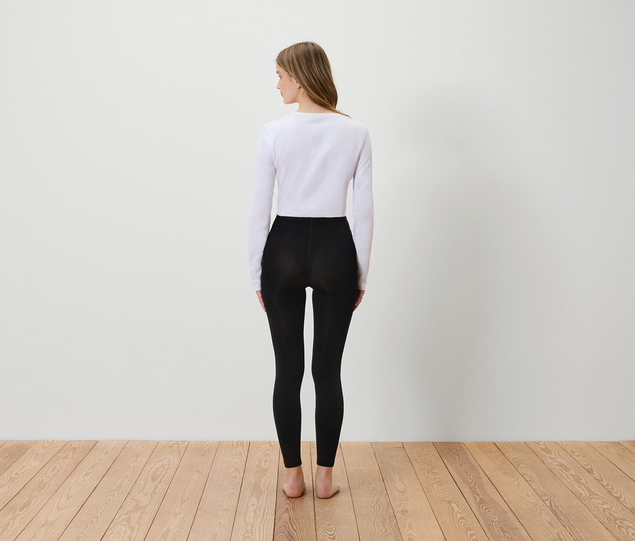 Frau steht mit dem Rücken zur Kamera, trägt ein weißes Langarmshirt und schwarze Leggings.