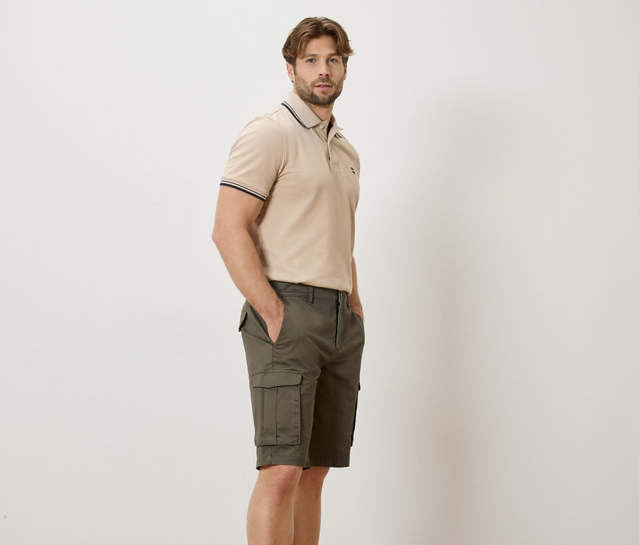 Mann steht seitlich in beigem Poloshirt und grünen Cargo-Shorts vor einem hellen Hintergrund.