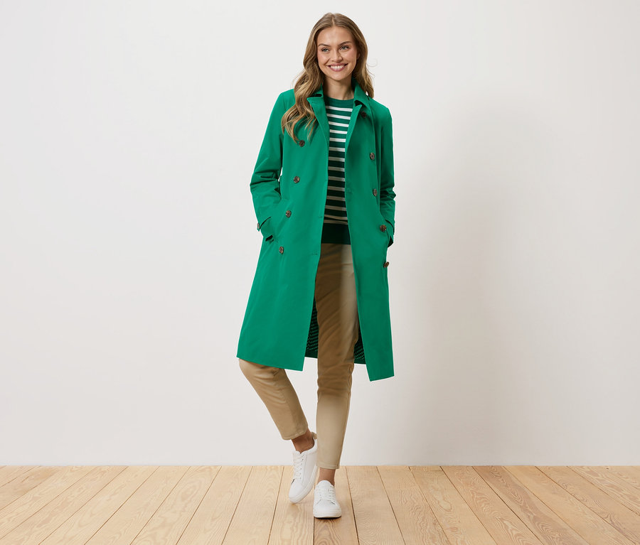 Frau mit grünem Mantel, gestreiftem Pullover, beigen Hosen und weißen Turnschuhen steht vor einer weißen Wand.