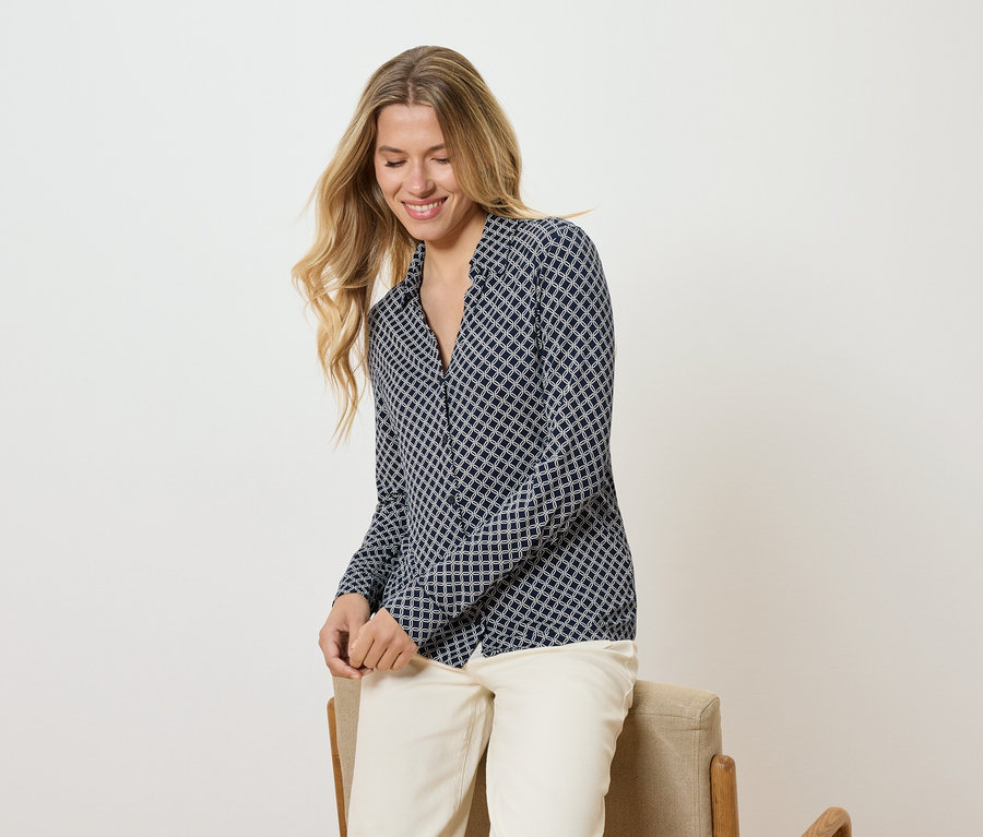 Frau mit langen blonden Haaren, die eine gemusterte Bluse und helle Hosen trägt, sitzt auf einem Stuhl und lächelt.