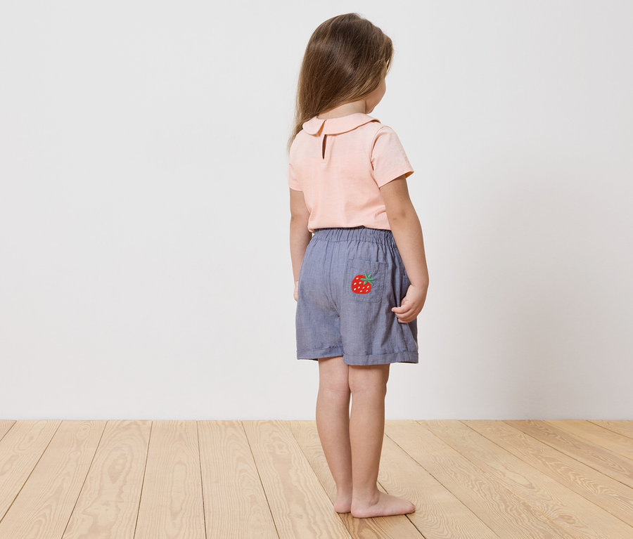 Mädchen von hinten, trägt ein rosa Hemd und blaue Shorts mit Erdbeerapplikation.