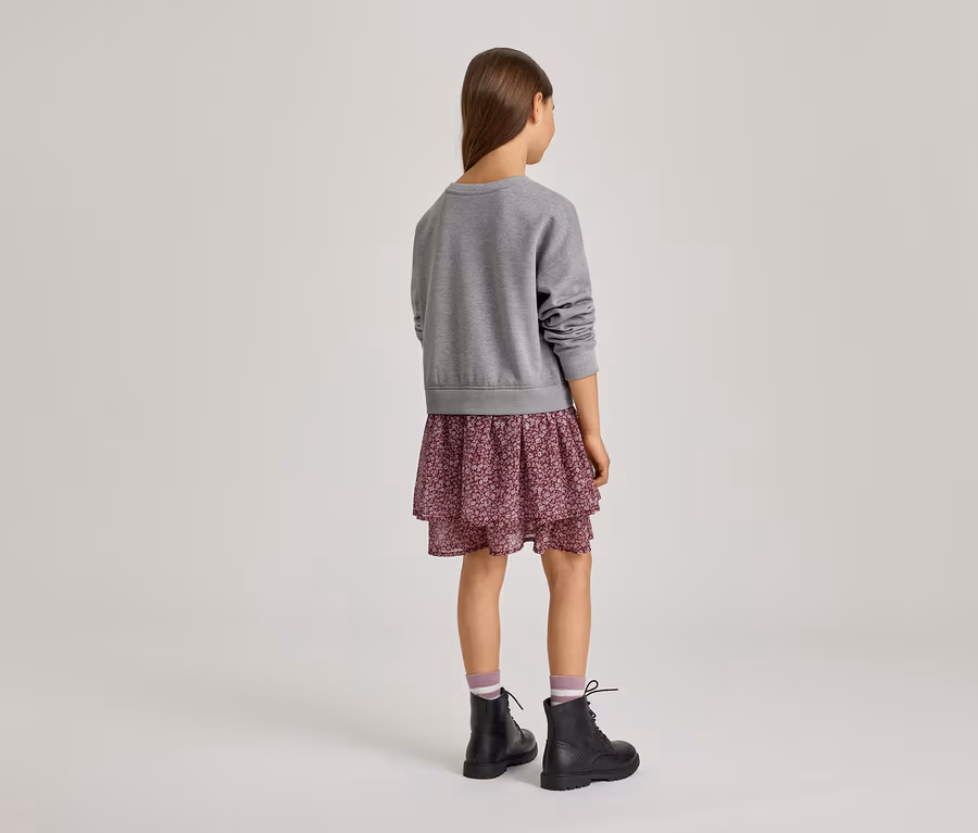 Mädchen von hinten, das einen grauen Pullover, einen gestuften Rock mit Blumenmuster, gestreifte Socken und schwarze Schnürstiefel trägt.