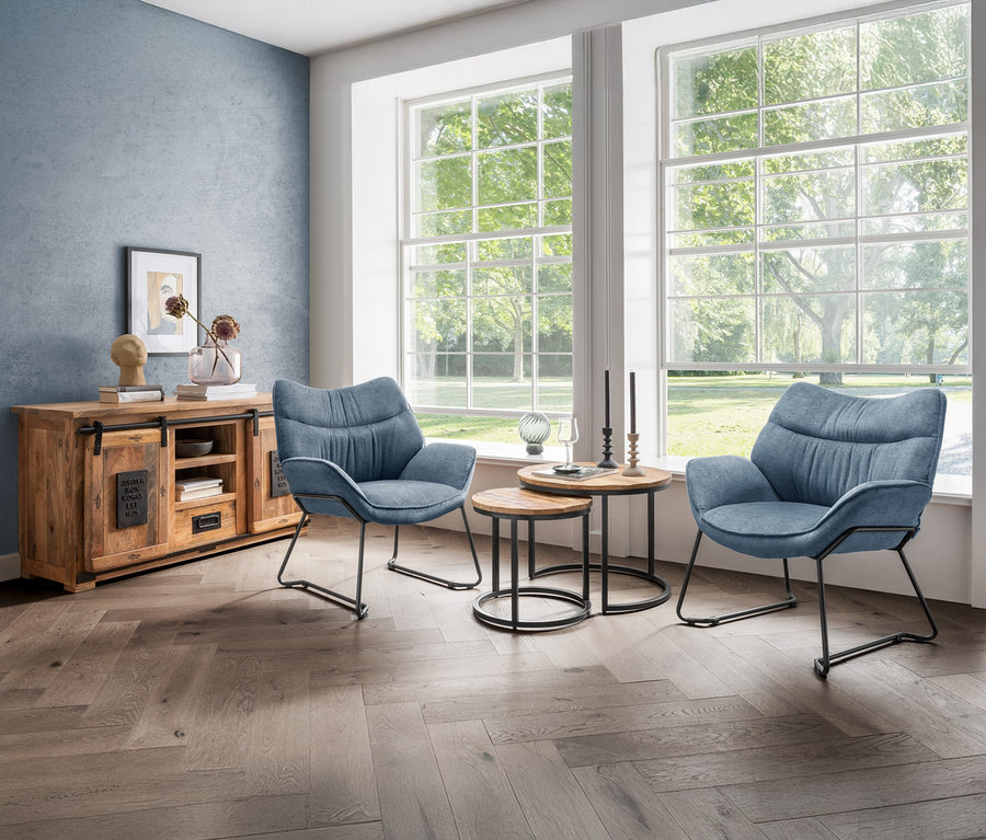 Wohnzimmer mit zwei blauen »Aliano« Sesseln von DeVries, Holztischchen und Kommode vor Fenstern.