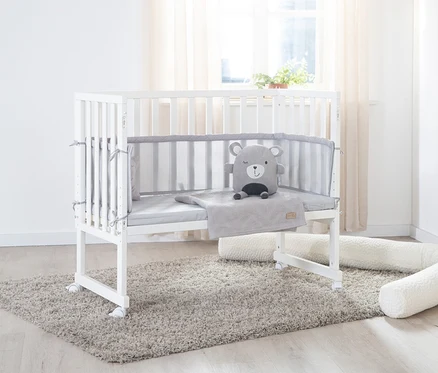 Weißes roba 3-in-1-Stuben-Beistellbett mit grauer Polsterung und Bär-Stofftier auf einem grauen Teppich.