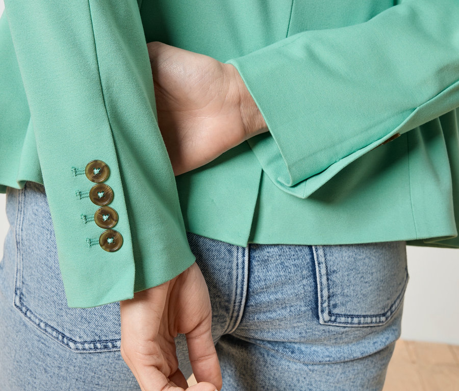 Nahaufnahme einer Person von hinten, die eine hellgrüne Jacke mit vier Knöpfen am Ärmel und eine Jeans trägt.