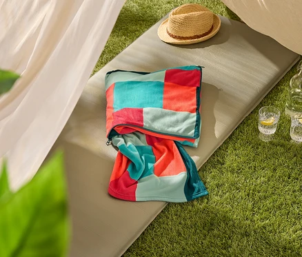Ein auf einer olivgrünen Isomatte liegendes, mehrfarbiges Strandtuch, daneben ein Strohhut und zwei Gläser Wasser mit Zitrone.