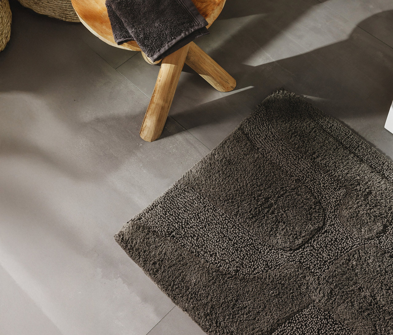 Eine graue benuta Badematte »Nehir« liegt auf einem Fliesenboden neben einem Hocker mit Handtüchern.