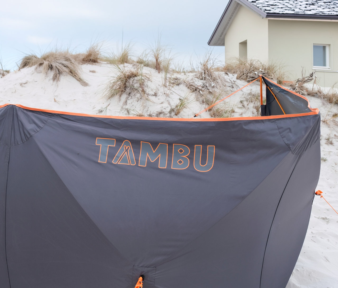 Grauer Windschutz mit orangefarbenen Akzenten und dem Schriftzug "TAMBU" auf Sand mit Gras und einem Haus im Hintergrund.