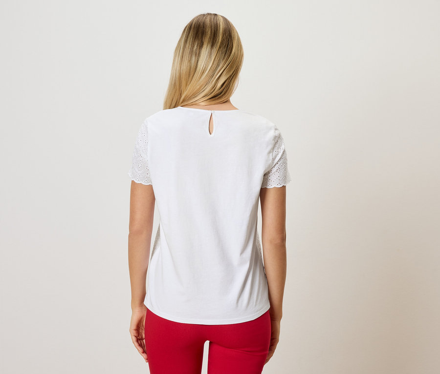 Rückansicht einer Frau mit blonden Haaren, die eine weiße Bluse mit Lochstickerei an den Ärmeln und eine rote Hose trägt.