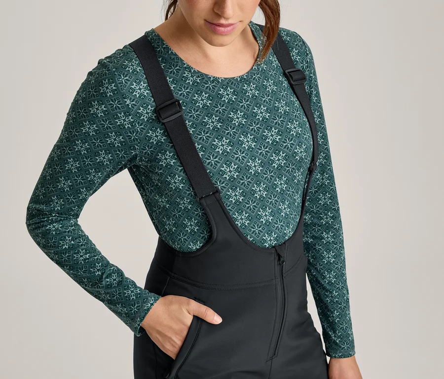 Frau trägt ein grünes Langarmshirt mit geometrischem Muster und eine schwarze Skihose mit Hosenträgern, ihre Hand steckt in einer Tasche.
