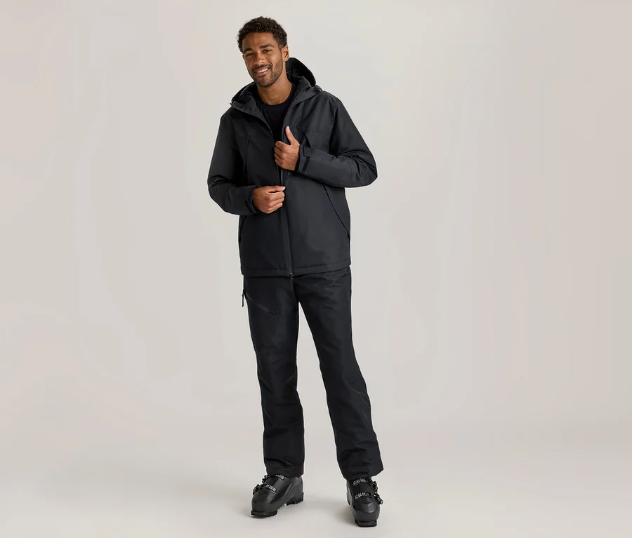Ein lächelnder Mann mit dunkler Haut und lockigem Haar steht da und richtet seine schwarze Skijacke. Er trägt eine schwarze Skijacke, schwarze Skihose und schwarze Skischuhe.