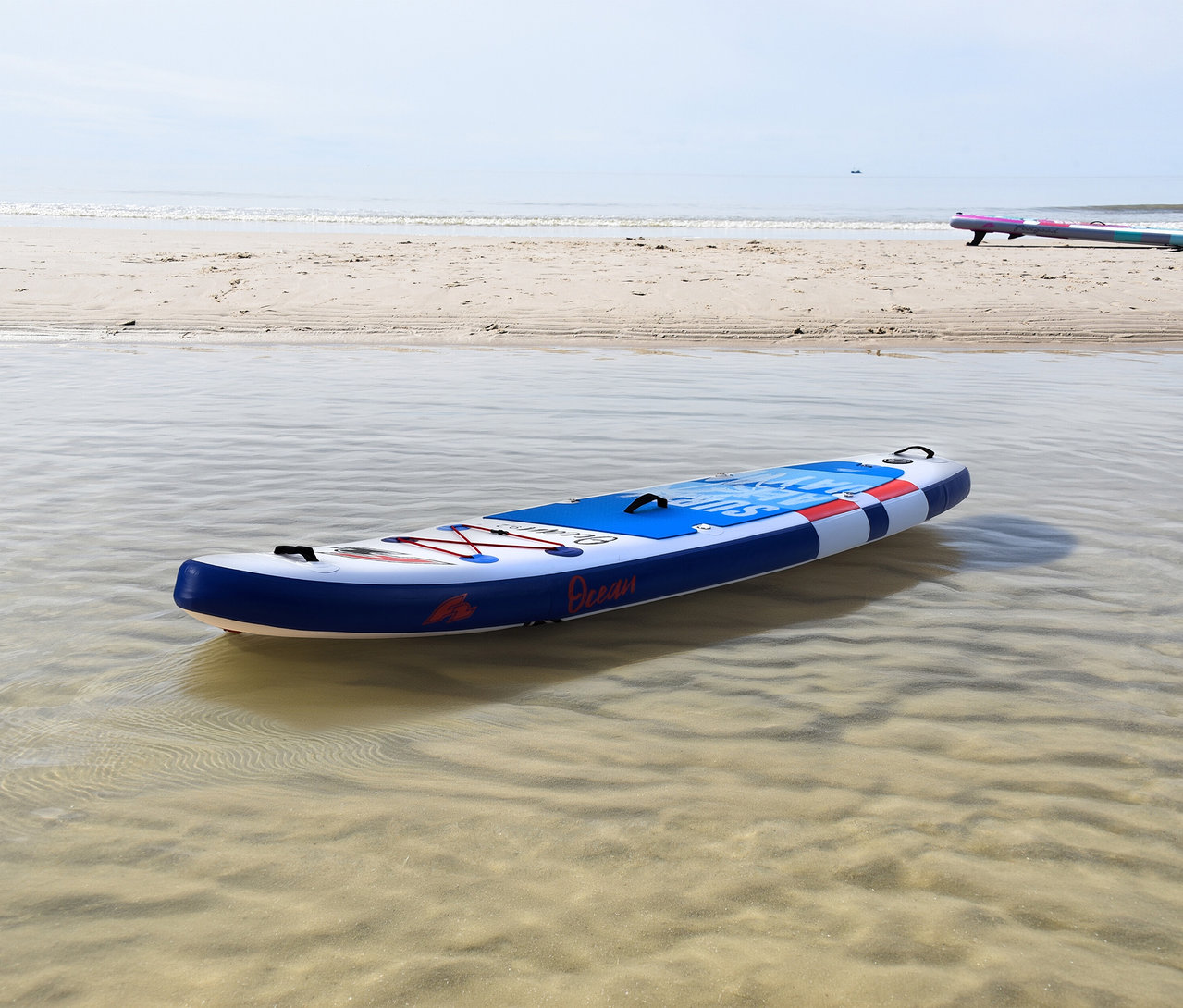Ein blau-weiß-rotes F2 SUP Board »Ocean Boy« schwimmt in flachem, klarem Wasser nahe einem Sandstrand. Im Hintergrund liegt ein weiteres SUP Board am Strand.