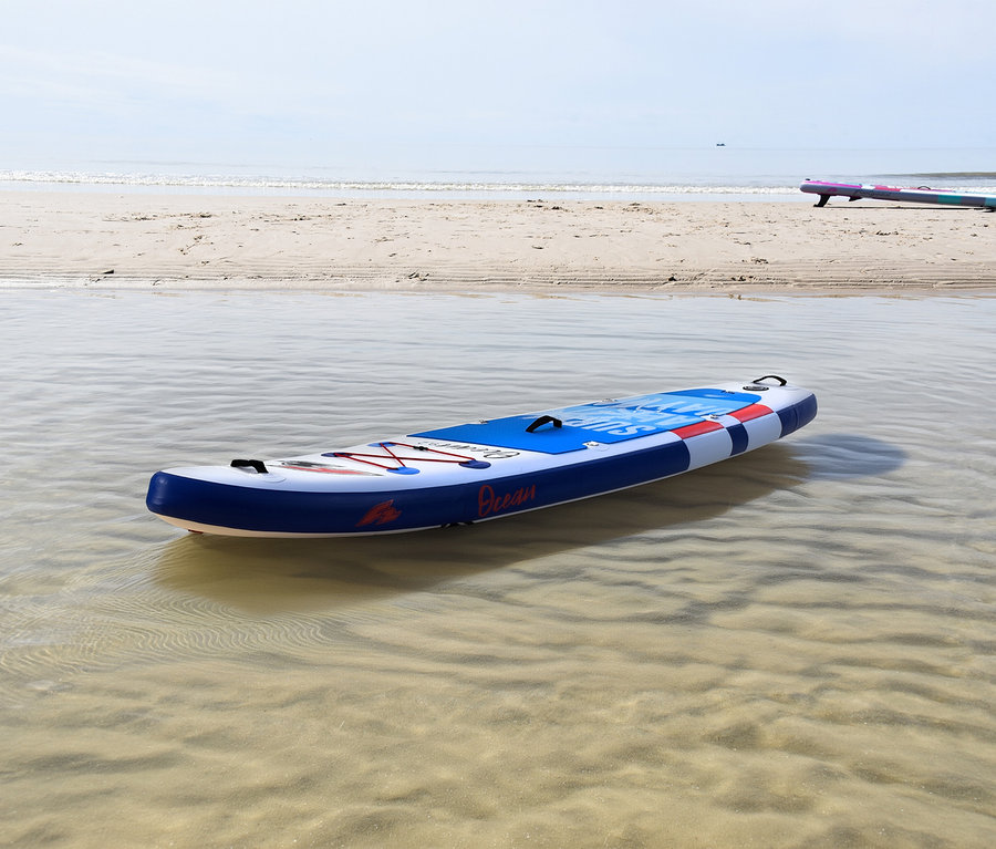 Ein blau-weiß-rotes F2 SUP Board »Ocean Boy« schwimmt in flachem, klarem Wasser nahe einem Sandstrand. Im Hintergrund liegt ein weiteres SUP Board am Strand.