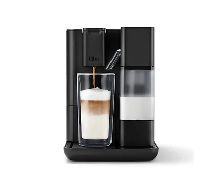 Eine schwarze Qbo MILK MASTER Velvet Black Kaffeemaschine brüht Kaffee in ein hohes Glas mit geschichtetem Milchkaffee, daneben ein Milchbehälter.