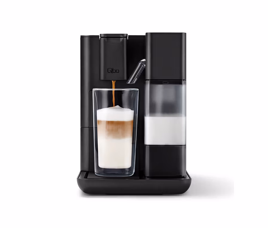 Eine schwarze Qbo MILK MASTER Velvet Black Kaffeemaschine brüht Kaffee in ein hohes Glas mit geschichtetem Milchkaffee, daneben ein Milchbehälter.