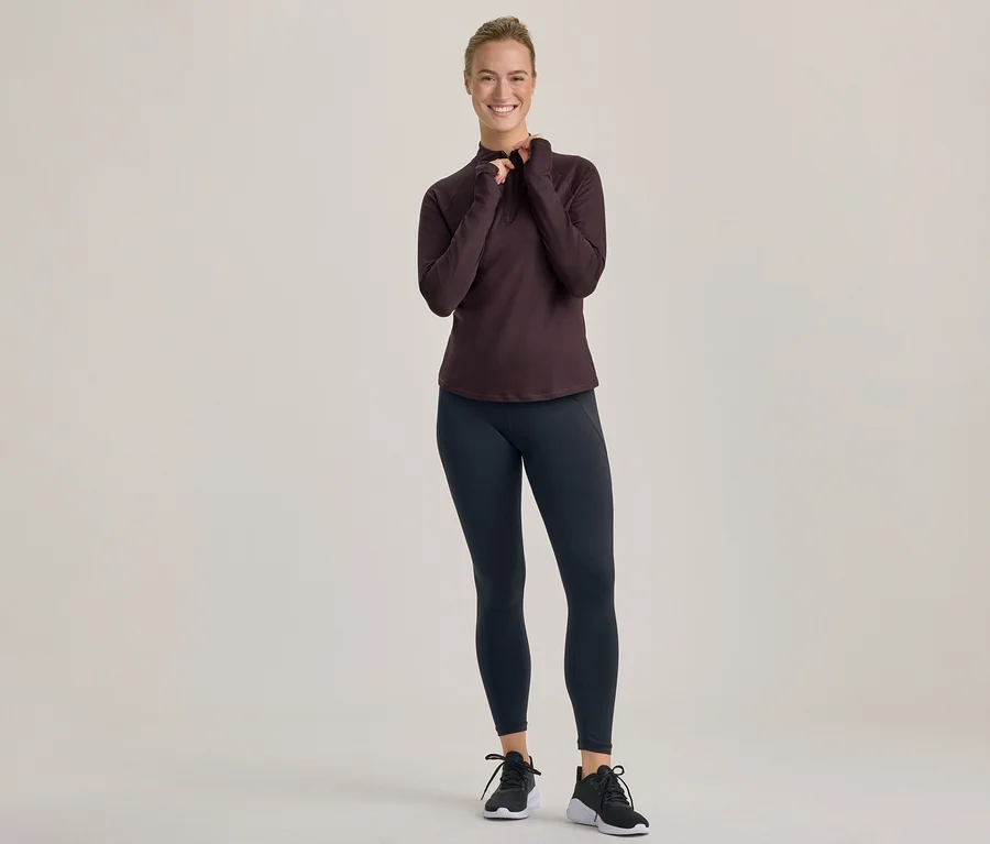 Lächelnde Frau in dunklem, langärmeligem Oberteil und schwarzen Leggings, die ihr Oberteil mit beiden Händen zurechtrückt.