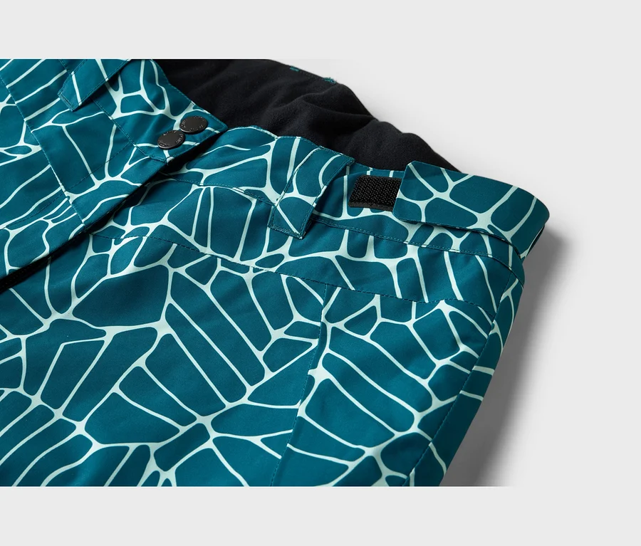 Nahaufnahme einer tealfarbenen Hose mit weißem geometrischem Muster, die den Bund mit zwei dunklen Knöpfen und einem schwarzen Klettverschluss sowie ein schwarzes Innenfutter zeigt.